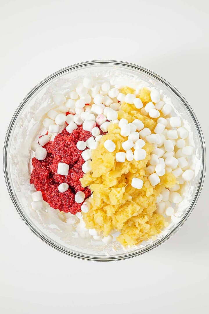Glass bowl with whipped cream, crushed pineapple, mini marshmallows, and a red fruit mixture on top.