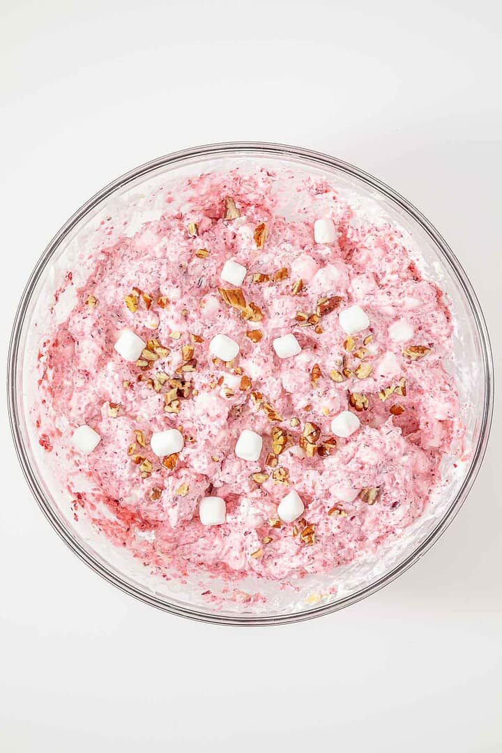 A glass bowl filled with pink whipped salad, topped with mini marshmallows and chopped pecans.