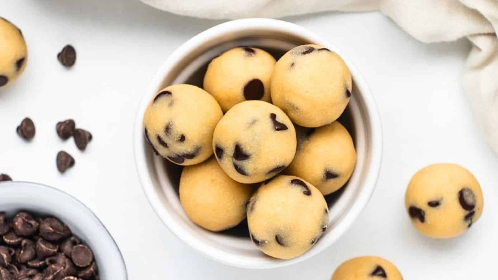 A bowl filled with round chocolate chip cookie dough balls on a white surface.