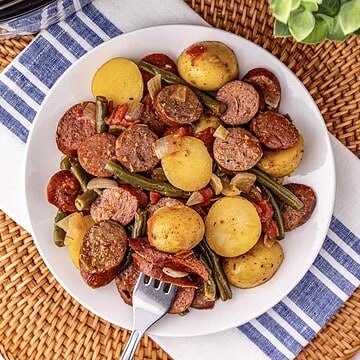 A plate of sausage, potatoes, green beans, and onions sits on a striped napkin with a fork on the plate.
