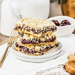Three crumbly cherry pie bars stacked on a white plate, with a fork and a bowl of cherries in the background—a delicious recipe perfect for dessert lovers.