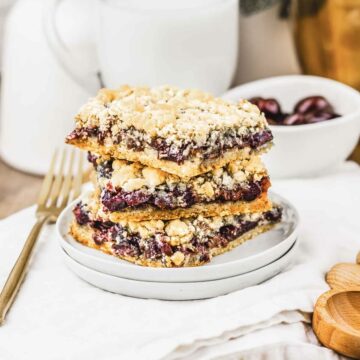 Three crumbly cherry pie bars stacked on a white plate, with a fork and a bowl of cherries in the background&mdash;a delicious recipe perfect for dessert lovers.