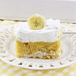 A slice of banana pudding poke cake topped with whipped cream and a banana slice on a white plate.