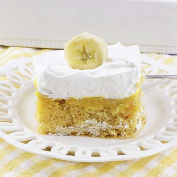 A slice of banana pudding poke cake topped with whipped cream and a banana slice on a white plate.