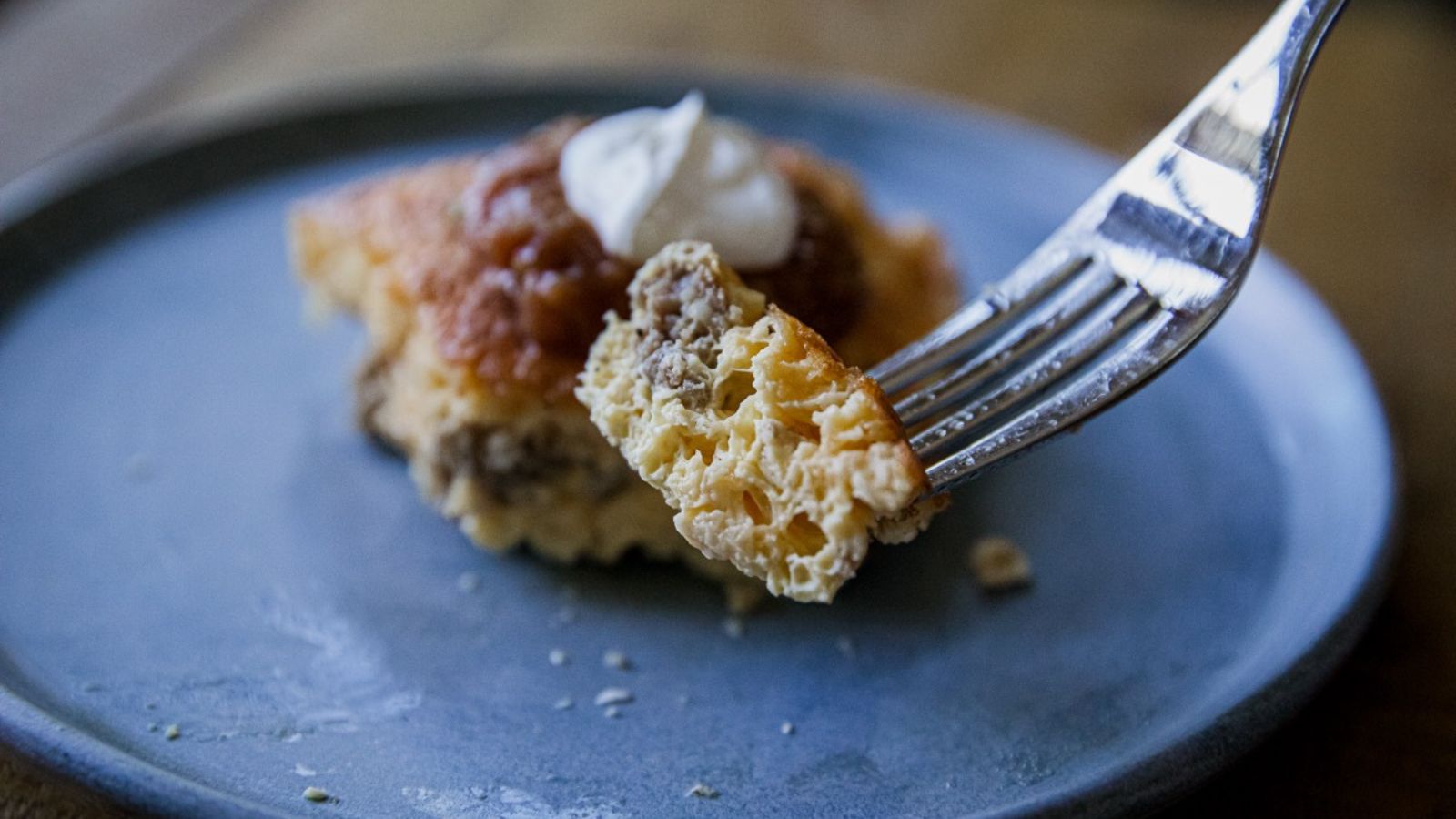 A fork holds a bite of cheesy casserole with ground meat, topped with sour cream, above a blue plate.