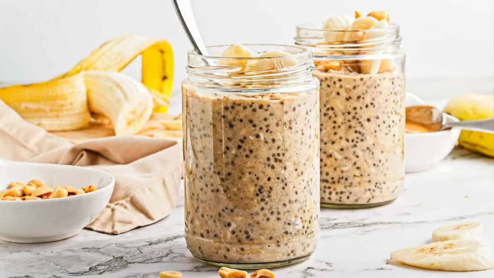 Two jars of chia seed overnight oats topped with banana slices and peanuts, with bananas and peanuts in the background.