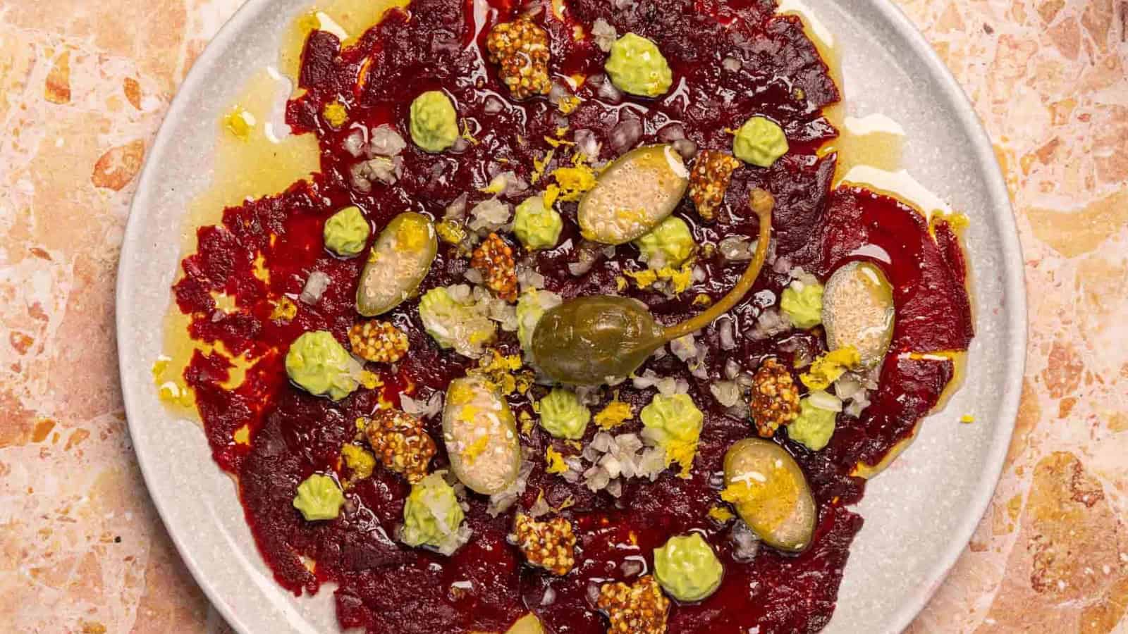 Colorful beet carpaccio topped with green cream, mustard seeds, capers, diced onions, and lemon zest on a plate.