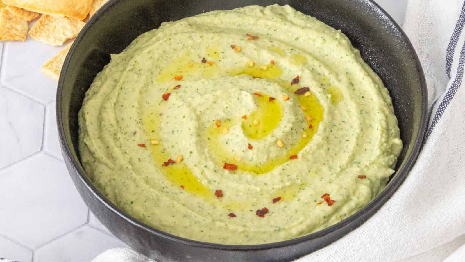 Creamy green hummus topped with olive oil and red pepper flakes in a black bowl, with pita chips nearby.