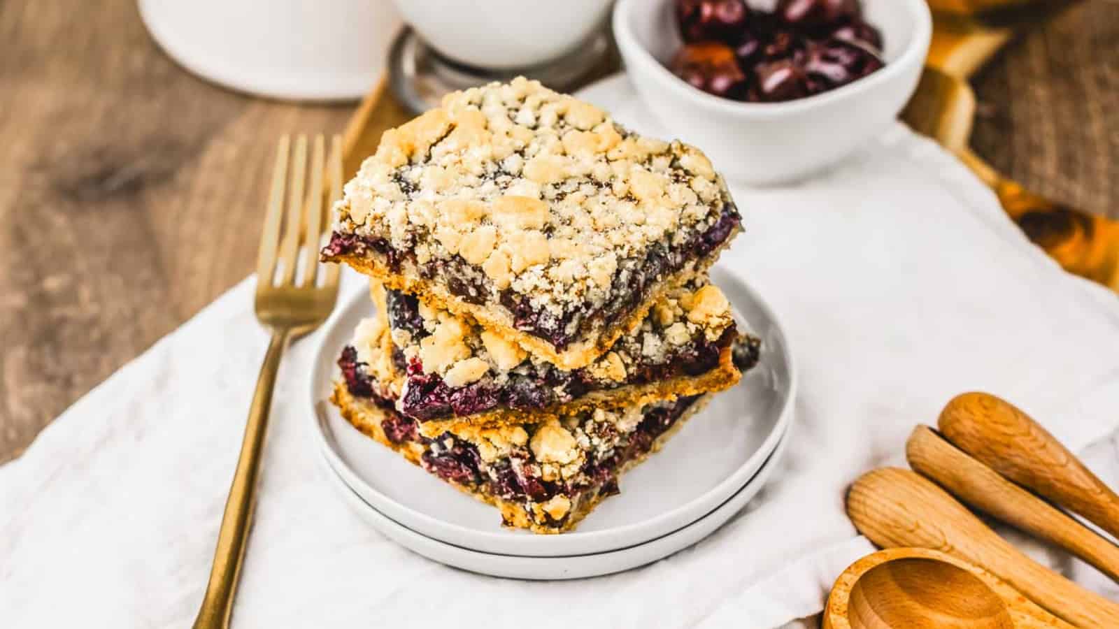 Three cherry crumble bars stacked on a white plate beside a gold fork and wooden measuring spoons.