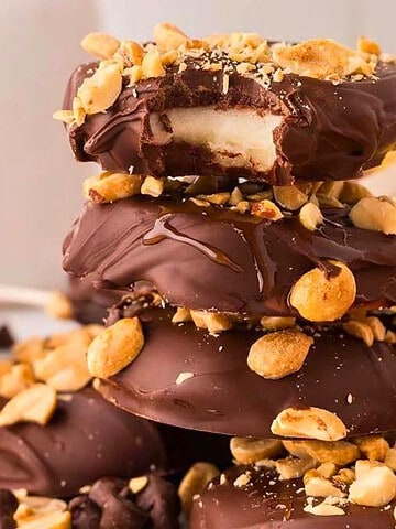 Three chocolate-covered apple slices on sticks, stacked and topped with chopped peanuts, surrounded by apples.