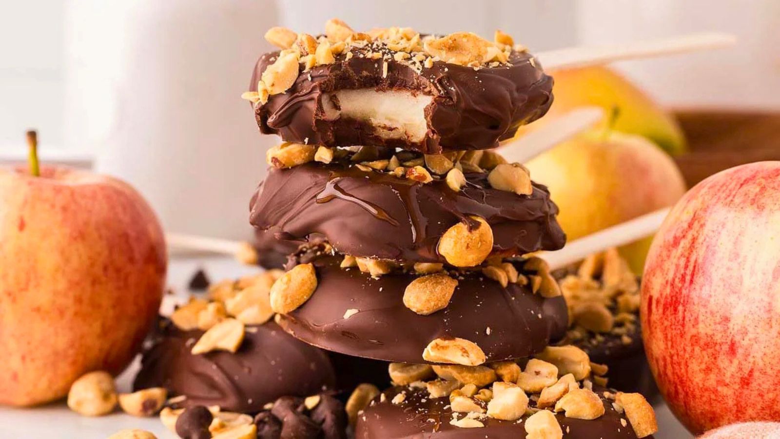 Three chocolate-covered apple slices on sticks, stacked and topped with chopped peanuts, surrounded by apples.