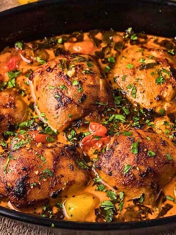 Roasted chicken thighs with herbs, tomatoes, and peppers in a black baking dish.