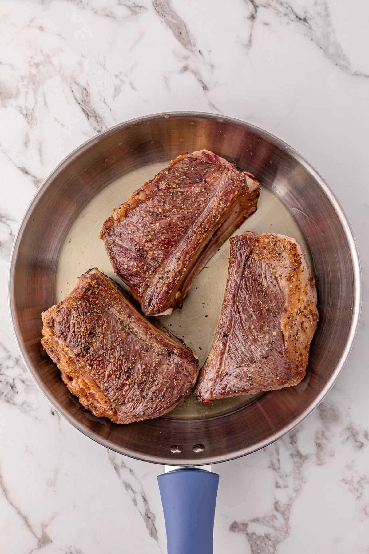Three seasoned beef short ribs searing in a stainless steel pan on a marble countertop, perfect for finishing in your slow cooker.