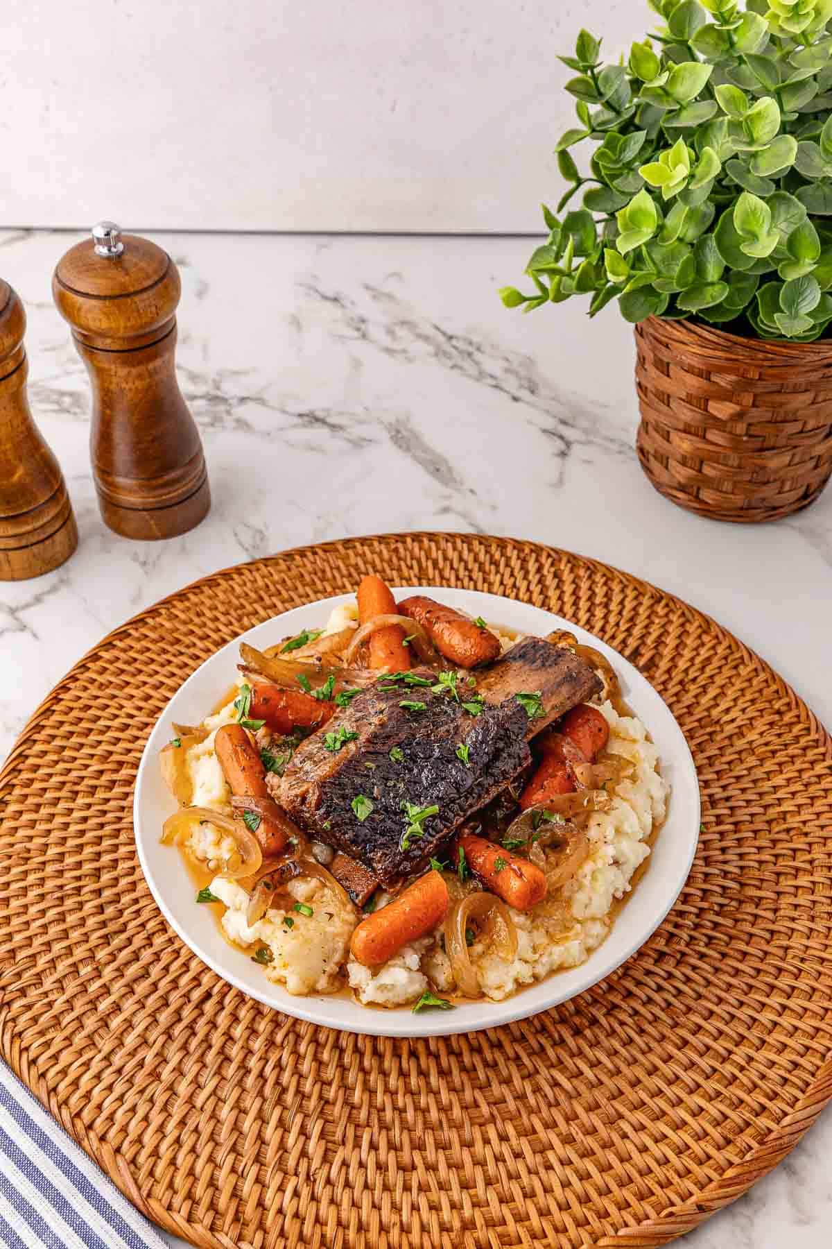 A plate of Slow Cooker Beef pot roast with carrots and potatoes served over mashed potatoes, on a woven placemat with a plant nearby.