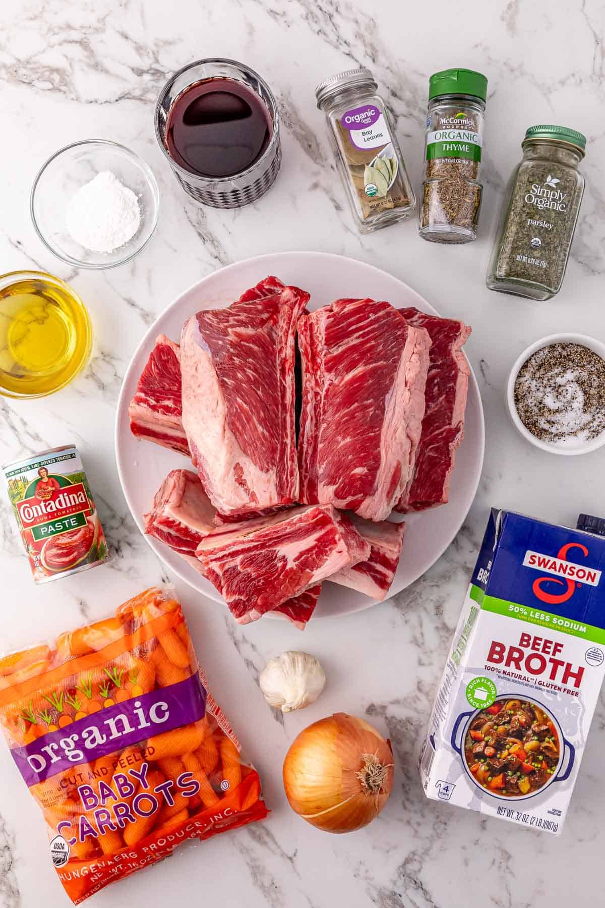 Raw beef short ribs on a plate, surrounded by beef broth, carrots, onion, seasonings, tomato paste, and red wine—perfect ingredients for tender slow cooker beef.