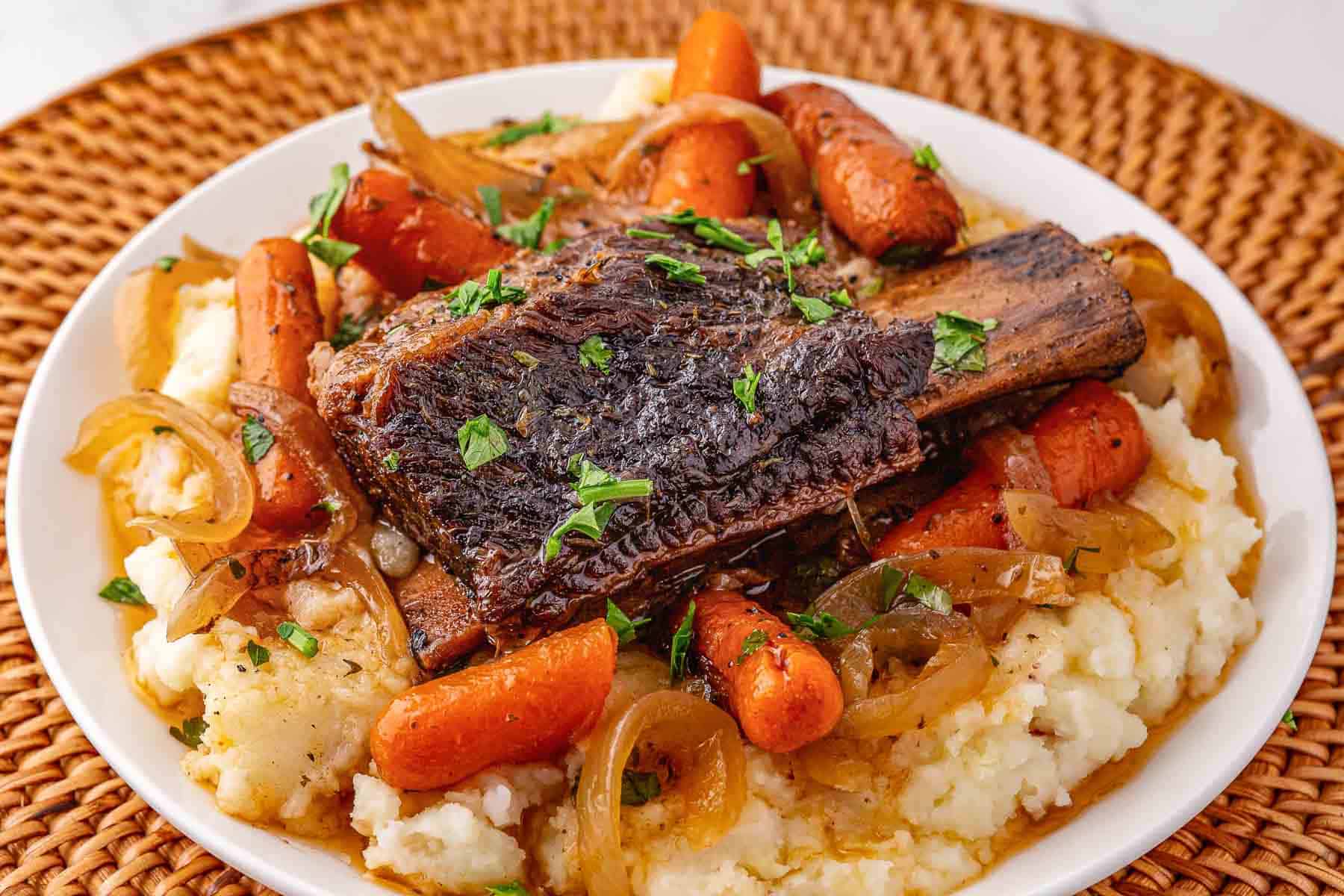 Slow cooker beef short ribs braised with carrots and onions, served over mashed potatoes and garnished with chopped parsley.