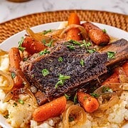 A plate of slow cooker beef short ribs with carrots, onions, and mashed potatoes, garnished with chopped herbs.
