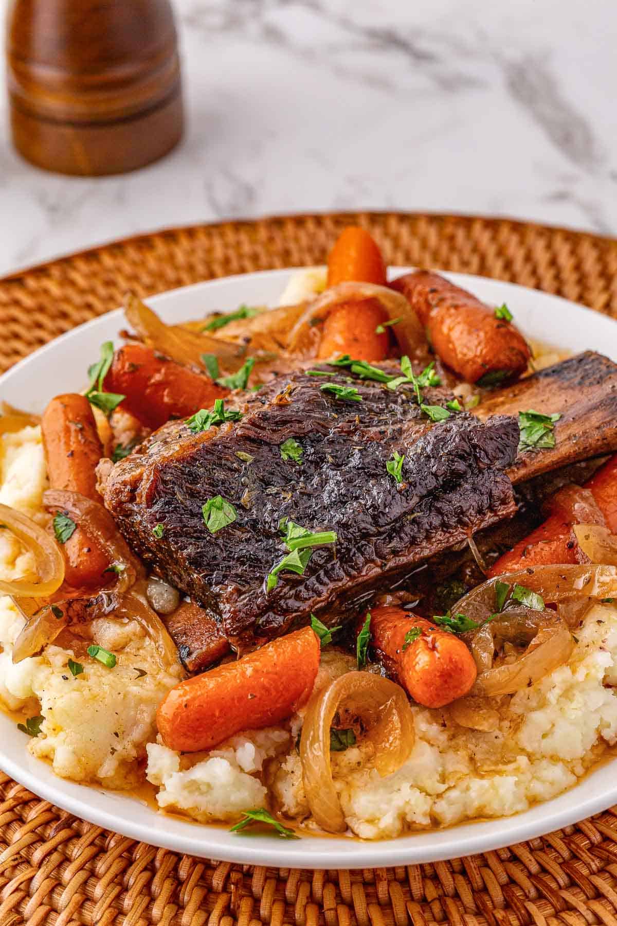 A plate of slow cooker beef short ribs with carrots, onions, and mashed potatoes, garnished with chopped herbs.