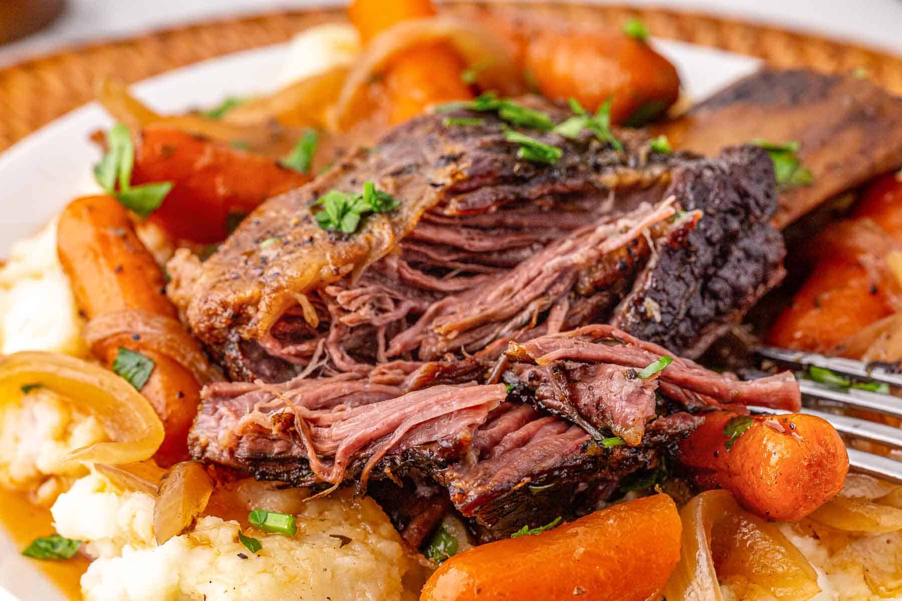 Tender beef short ribs with carrots, onions, and mashed potatoes, garnished with fresh herbs on a white plate.