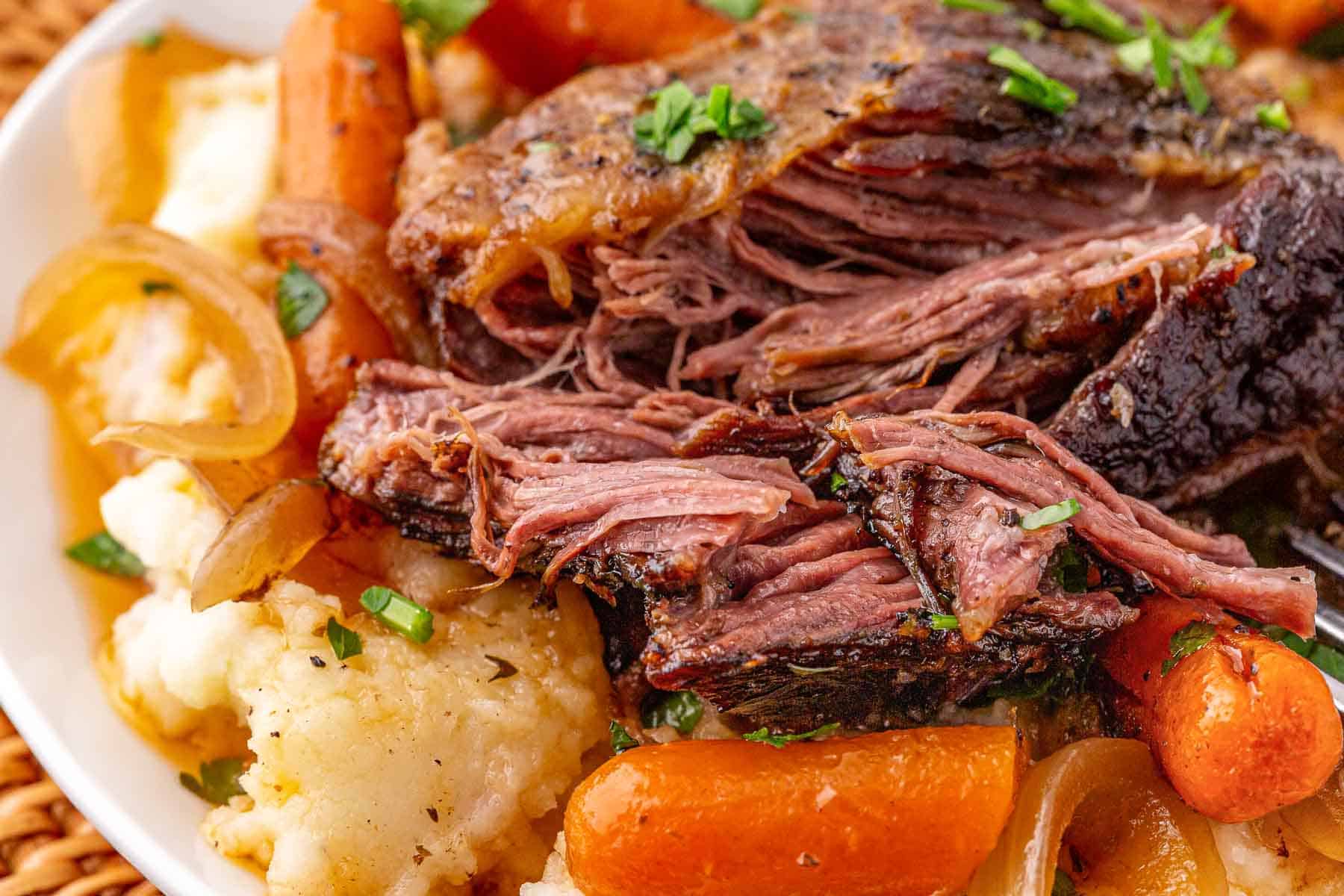 Tender pot roast with carrots, onions, and mashed potatoes, garnished with fresh herbs on a white plate—perfect comfort food inspired by classic Slow Cooker Beef.