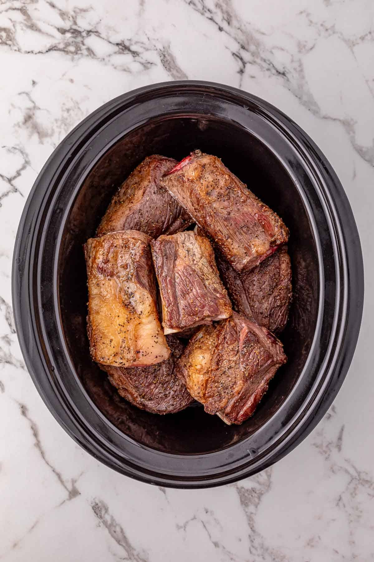 Beef short ribs, perfectly browned, rest in a black slow cooker on a white marble surface.
