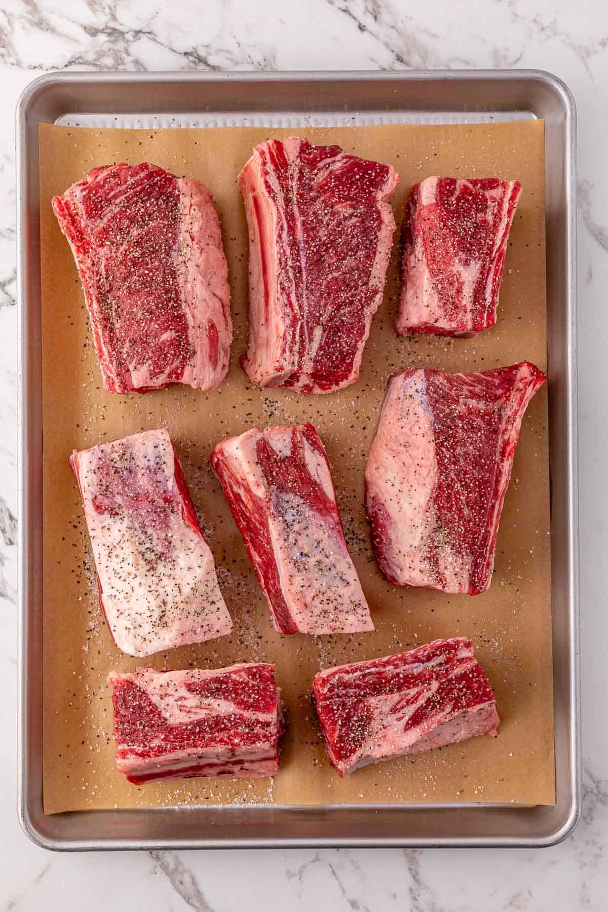 Eight seasoned beef short ribs on a parchment-lined baking sheet, ready for the slow cooker or oven, set atop a marble surface.