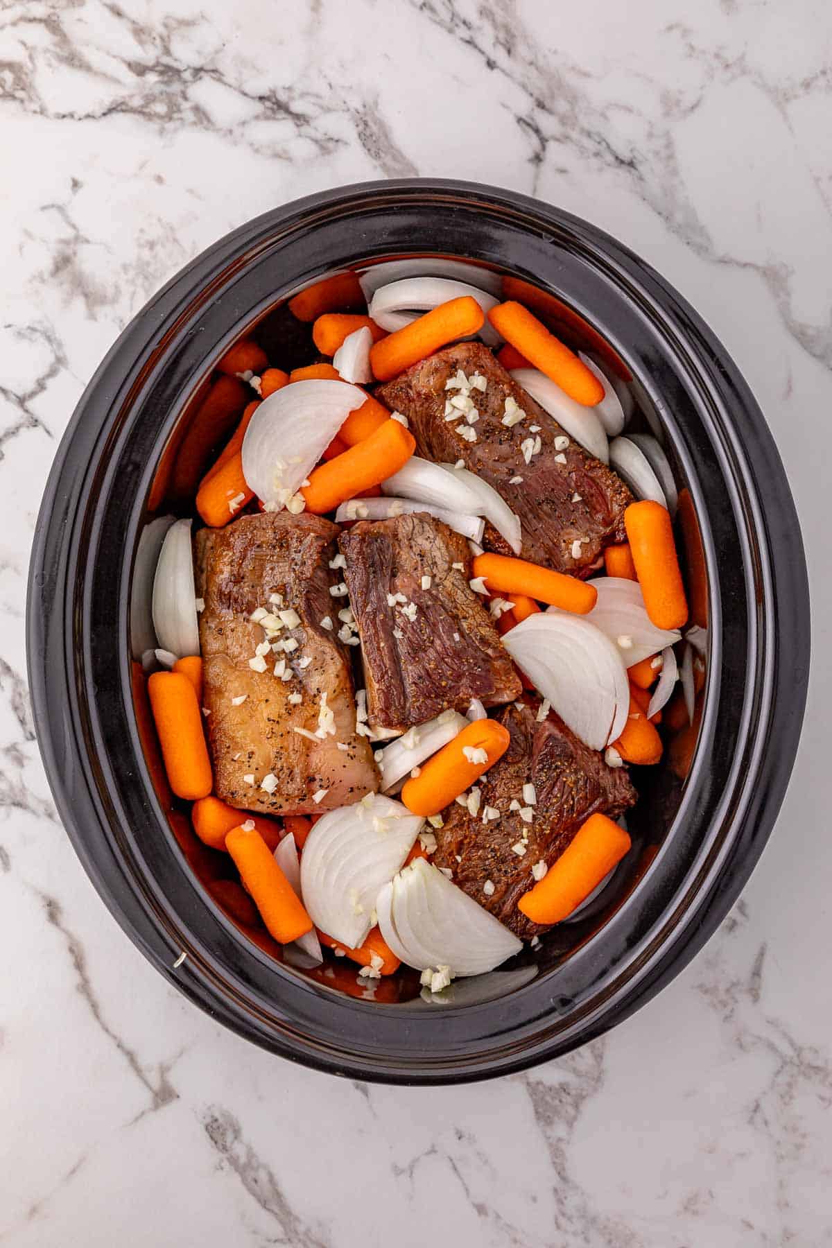 Beef short ribs, baby carrots, chopped onions, and minced garlic are ready in the slow cooker on a marble countertop—perfect for a hearty slow cooker beef dinner.