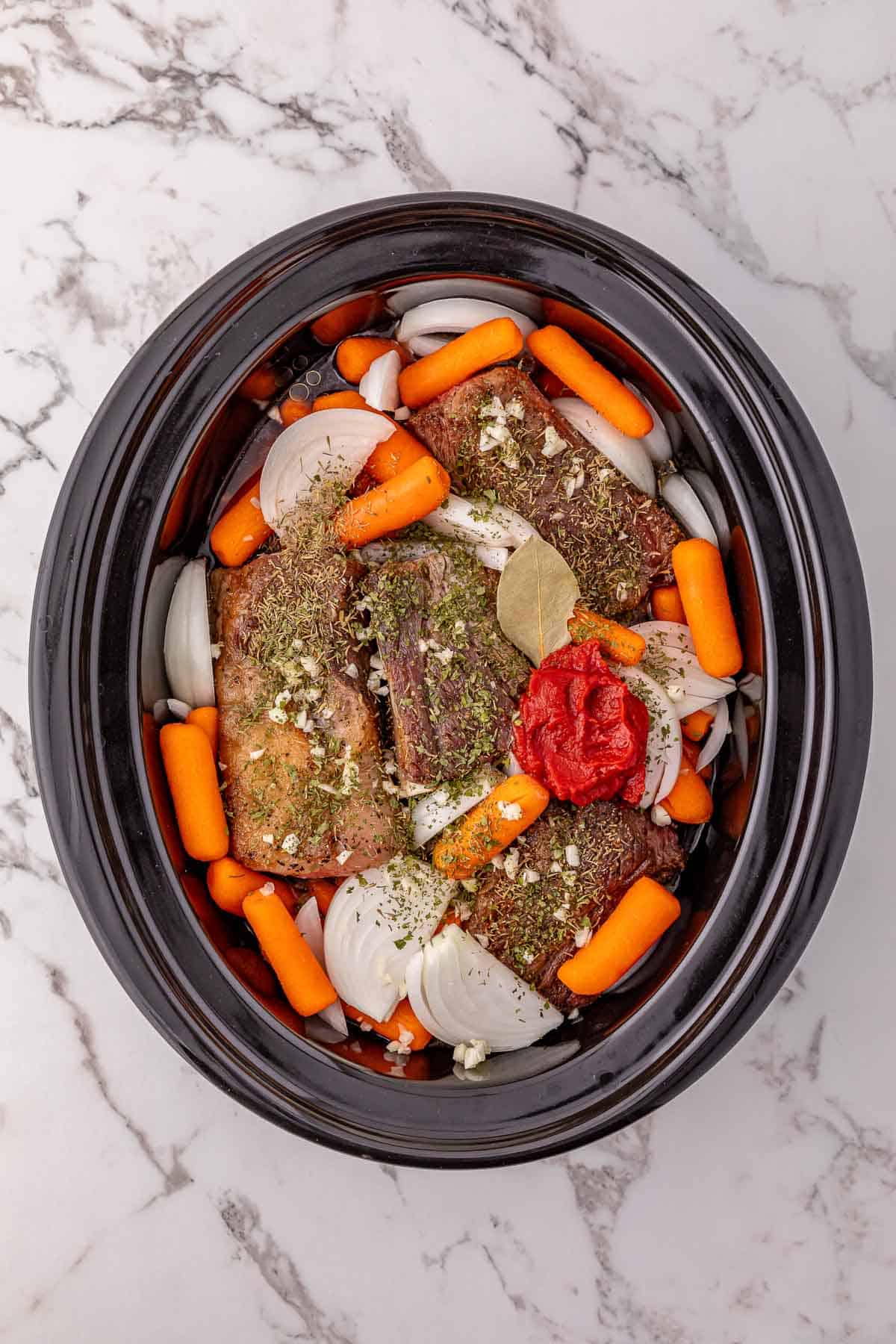 Beef short ribs, sliced onions, carrots, herbs, and tomato paste in a slow cooker on a marble countertop.