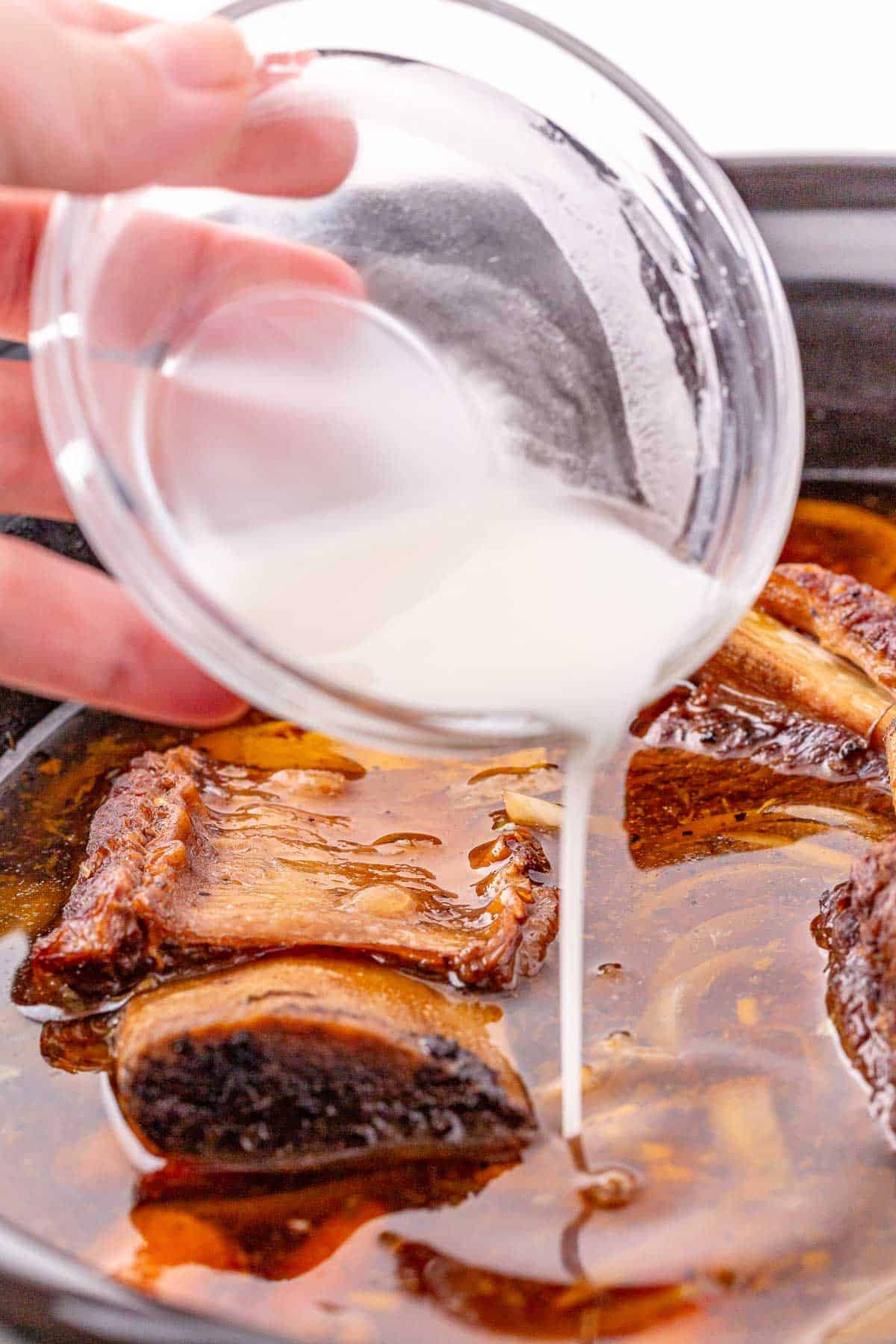 A hand pours a cloudy liquid from a small bowl into a pot of slow cooker beef short ribs simmering in savory broth.