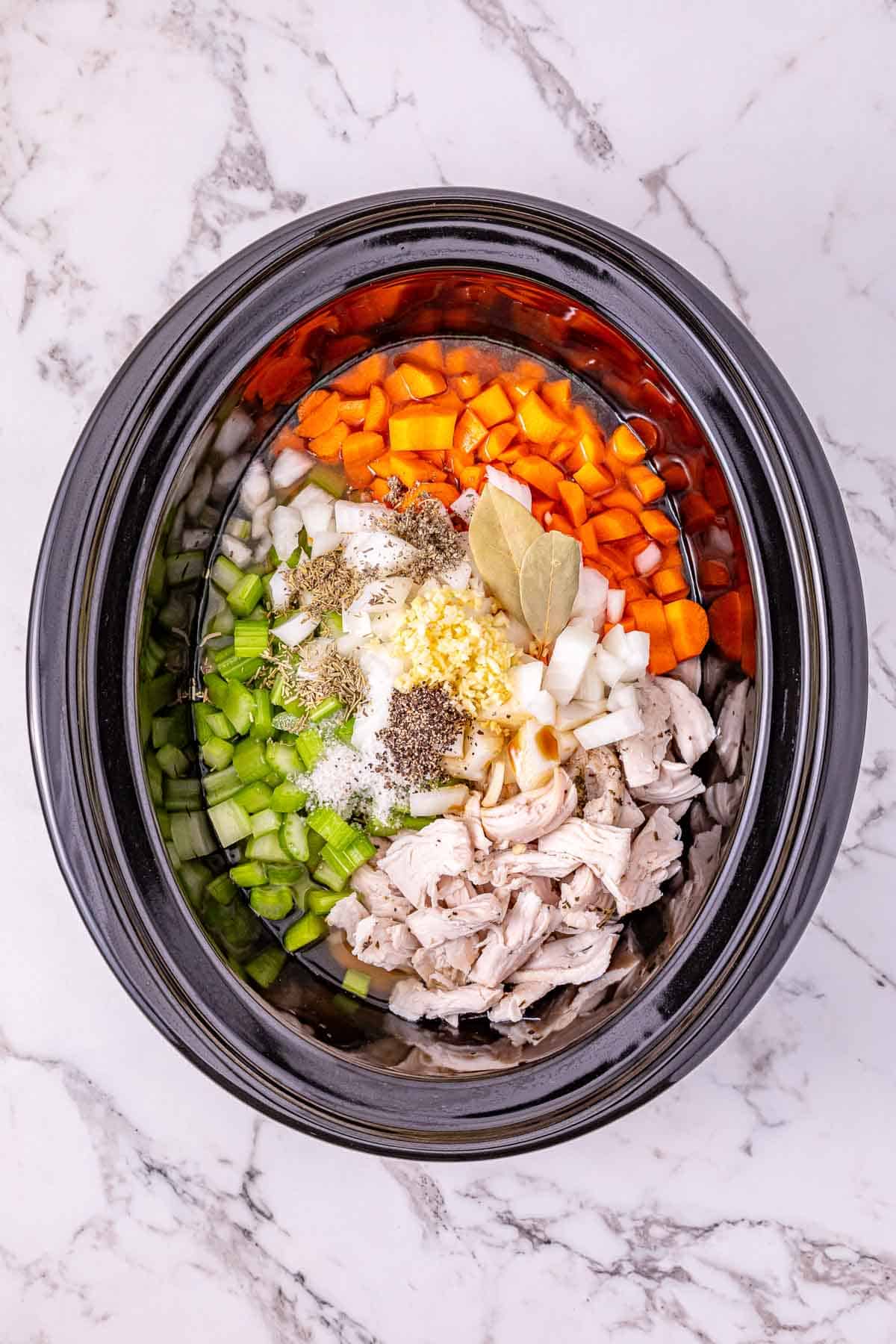 Slow cooker filled with chopped carrots, celery, onion, leftover turkey, rice, spices, and a bay leaf—everything you need for a comforting leftover turkey soup ready to cook.