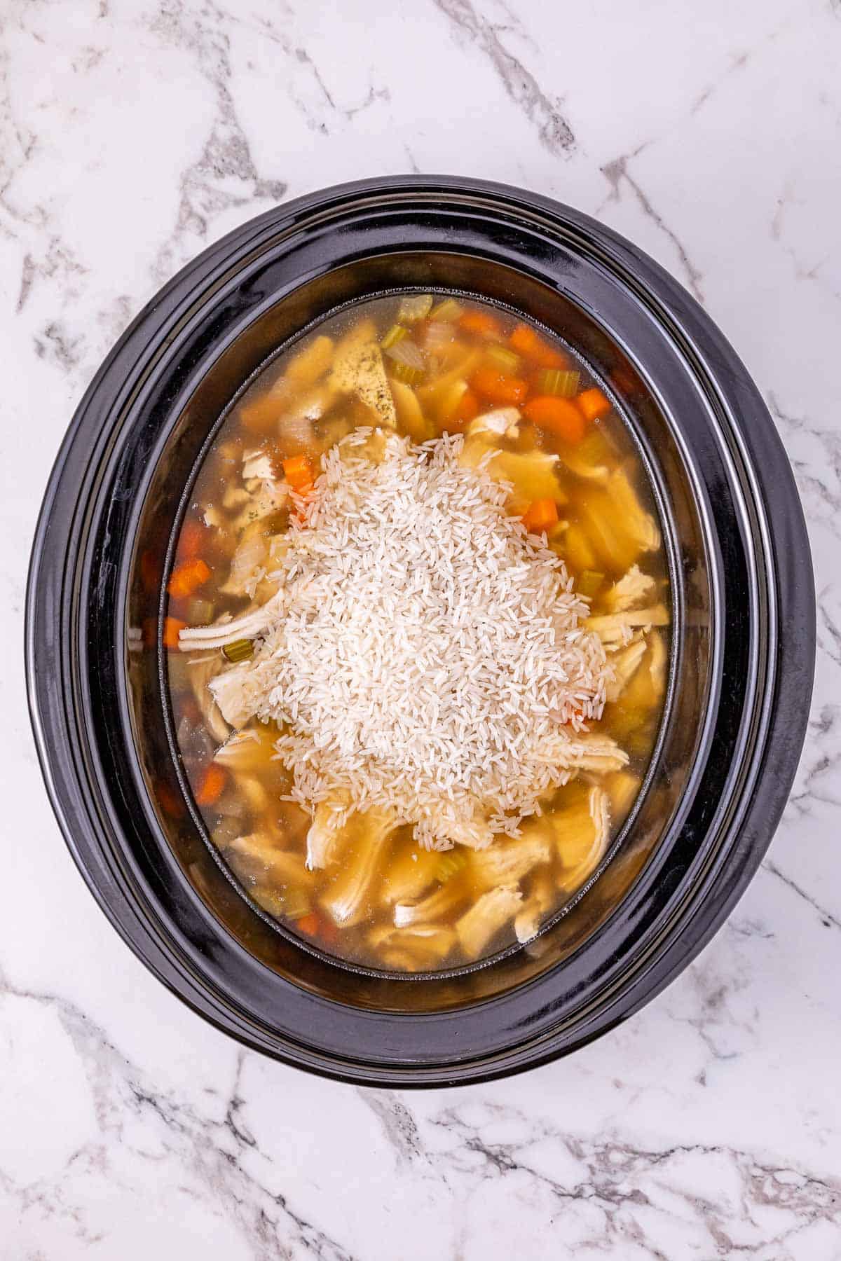 Overhead view of uncooked rice added to a slow cooker with leftover turkey and soup ingredients on a marble surface, perfect for making a hearty slow cooker turkey soup.