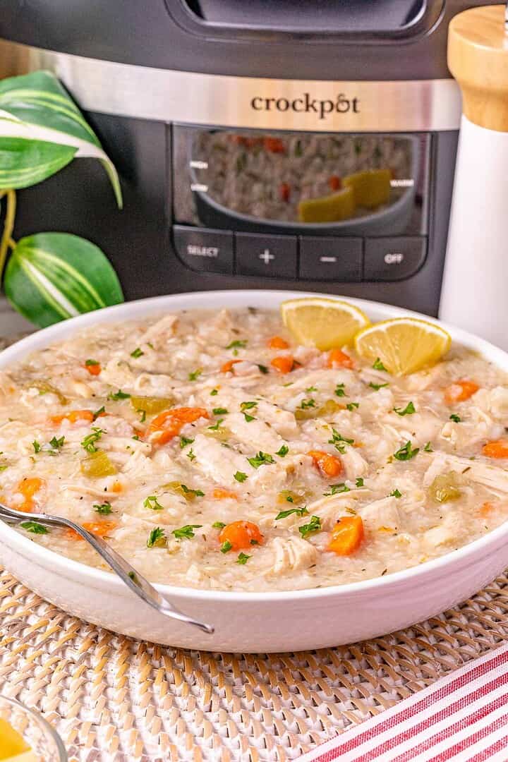 A bowl of creamy slow cooker turkey soup with rice and carrots, garnished with lemon wedges and parsley, in front of a Crockpot.