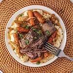 Plate of tender slow cooker beef pot roast with carrots, onions, and parsley on mashed potatoes, with a fork on the side.