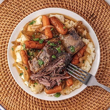 Plate of tender slow cooker beef pot roast with carrots, onions, and parsley on mashed potatoes, with a fork on the side.