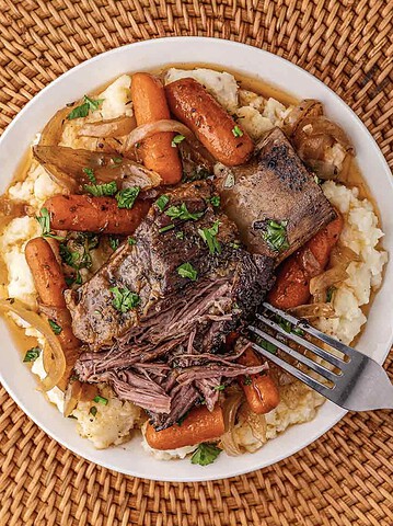 Plate of tender slow cooker beef pot roast with carrots, onions, and parsley on mashed potatoes, with a fork on the side.