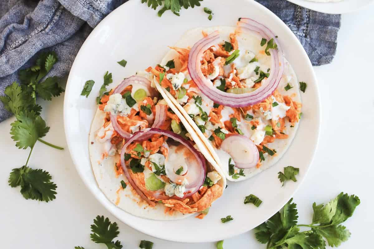 Two soft chicken tacos with shredded Buffalo chicken filling, sliced red onions, green onions, and cilantro on a white plate—perfect for those who love easy recipes.