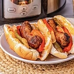 Two sausage sandwiches with peppers and onions on a white plate, in front of a kitchen appliance.