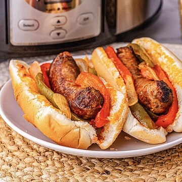 Two sausage sandwiches with peppers and onions on a white plate, in front of a kitchen appliance.