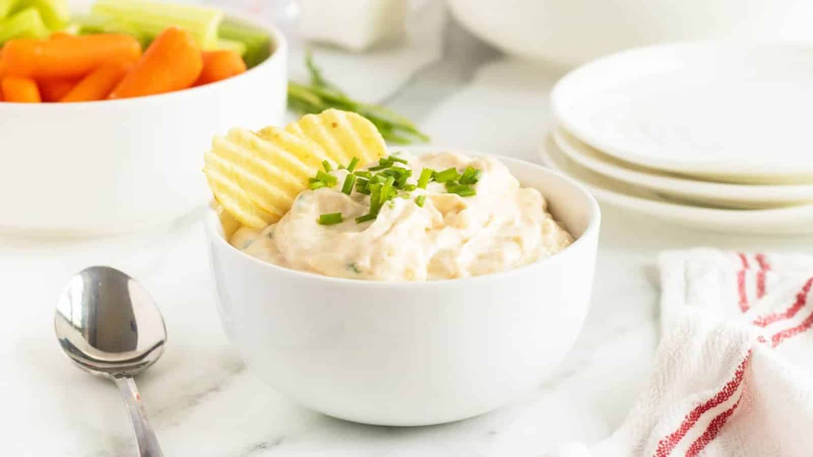 A bowl of creamy dip topped with chives and potato chips, with veggies and plates in the background.