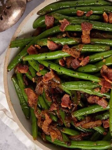 Green beans topped with crispy bacon pieces in a bowl, with a serving spoon and napkin beside it.