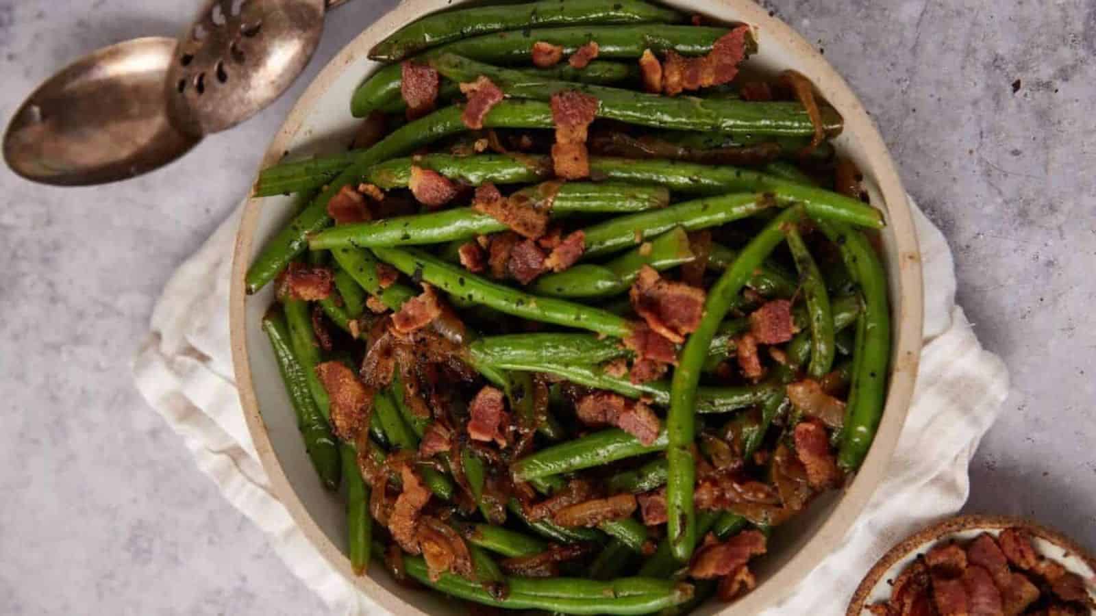 Green beans topped with crispy bacon pieces in a bowl, with a serving spoon and napkin beside it.