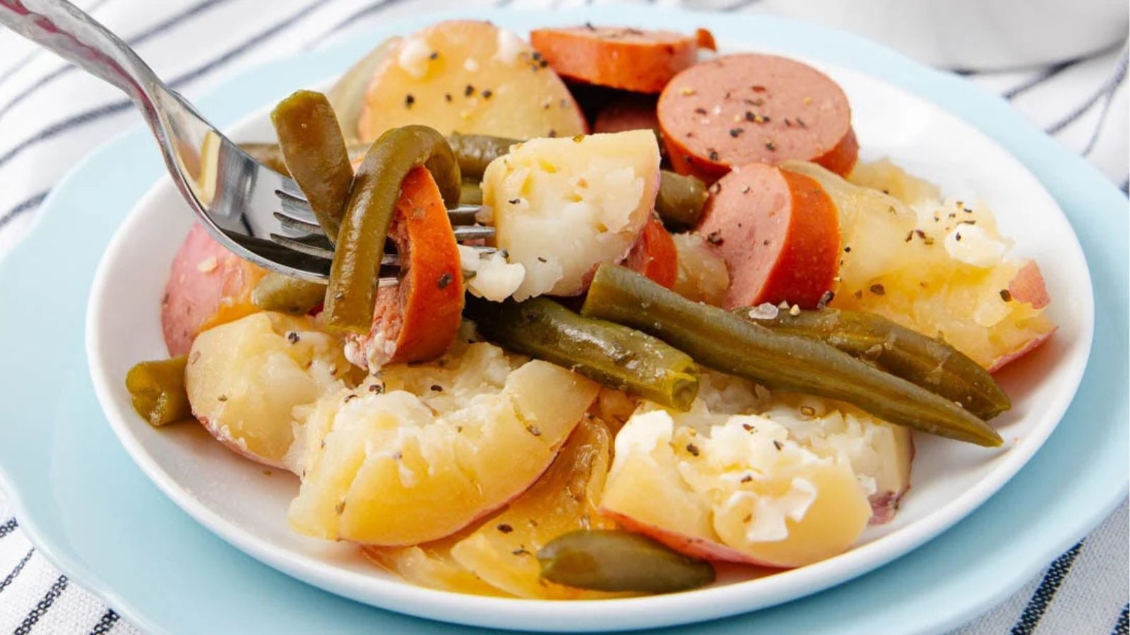 A plate of sausage, green beans, and potatoes with pepper, being picked up with a fork.
