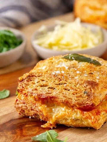 Golden, cheesy pastry squares with herbs on a wooden board, garnished with chives and cheese in bowls.