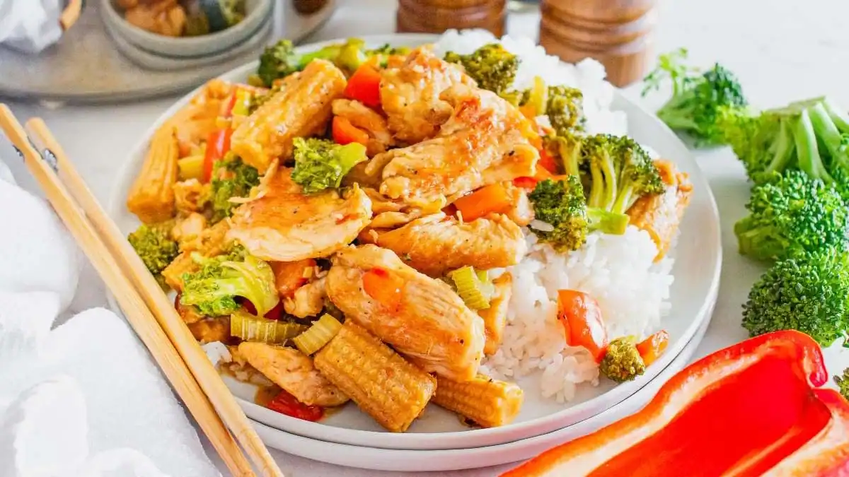 A plate of white rice topped with stir-fried chicken, baby corn, broccoli, and carrots, with chopsticks nearby.