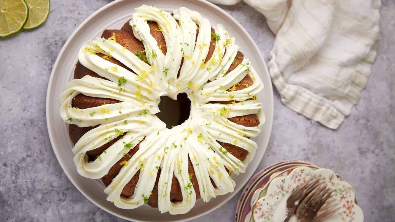 Bundt cake with white icing and citrus zest on top, served on a plate with limes and napkins nearby.
