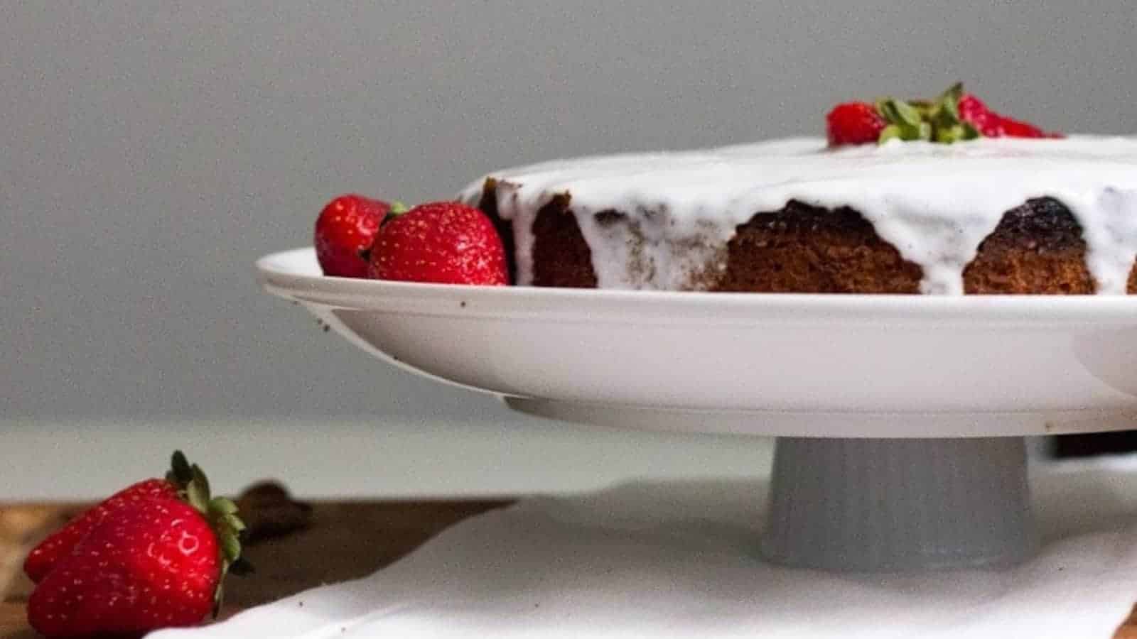 A frosted cake topped with strawberries sits on a white cake stand, surrounded by more strawberries—perfect inspiration for simple cakes or anyone searching for easy cakes to try in their favorite cake recipes.