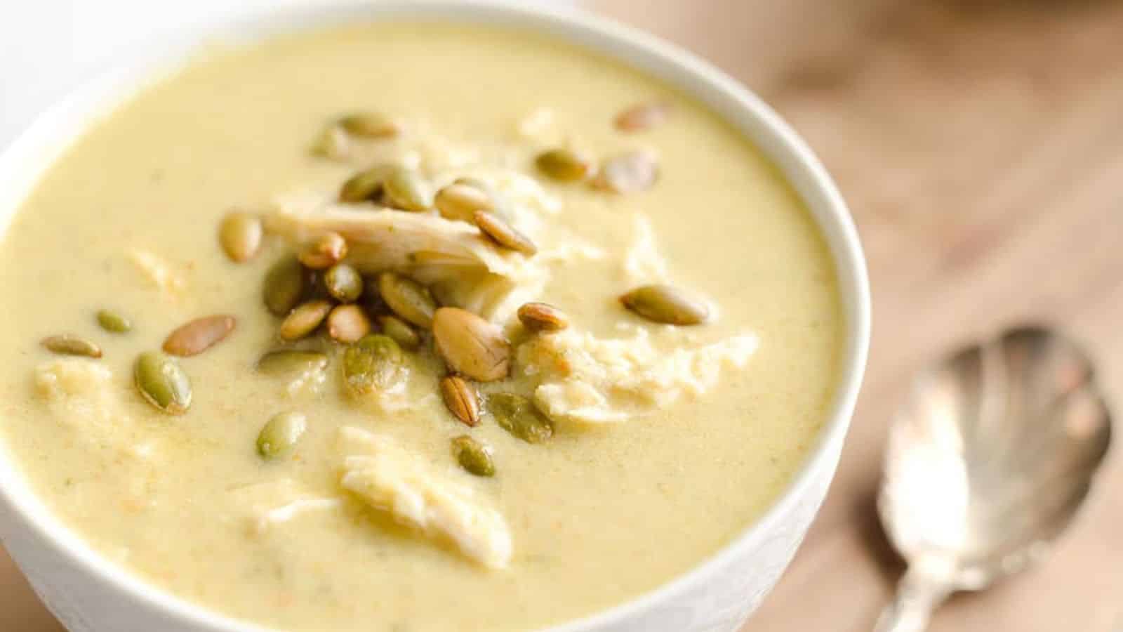 Bowl of creamy soup topped with shredded chicken and toasted pumpkin seeds, with a spoon beside it.