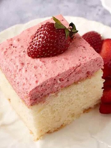 A slice of white cake with pink strawberry frosting, topped with a strawberry, and sliced strawberries on the side.
