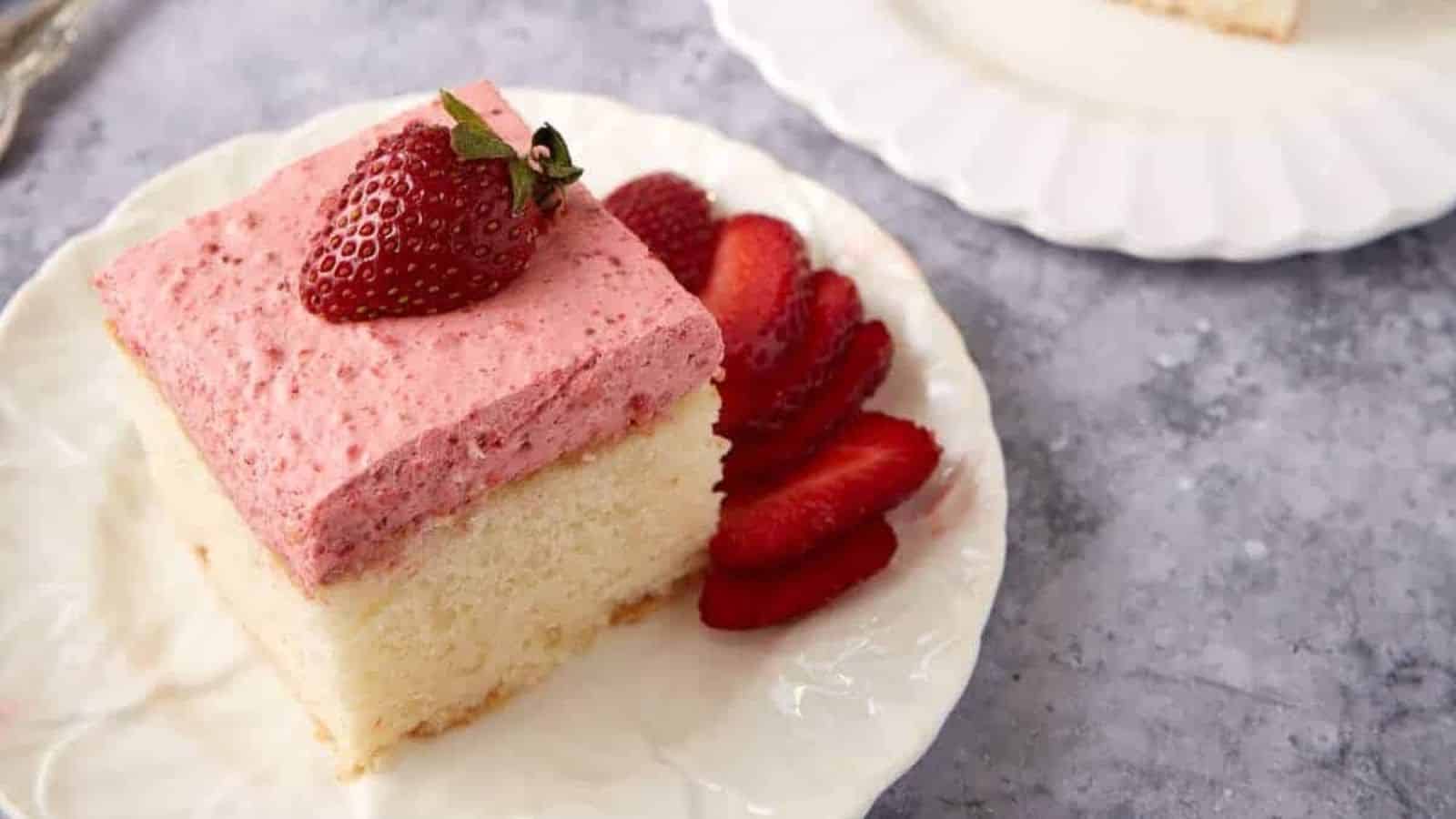 A slice of white cake with pink strawberry frosting, topped with a strawberry, and sliced strawberries on the side.