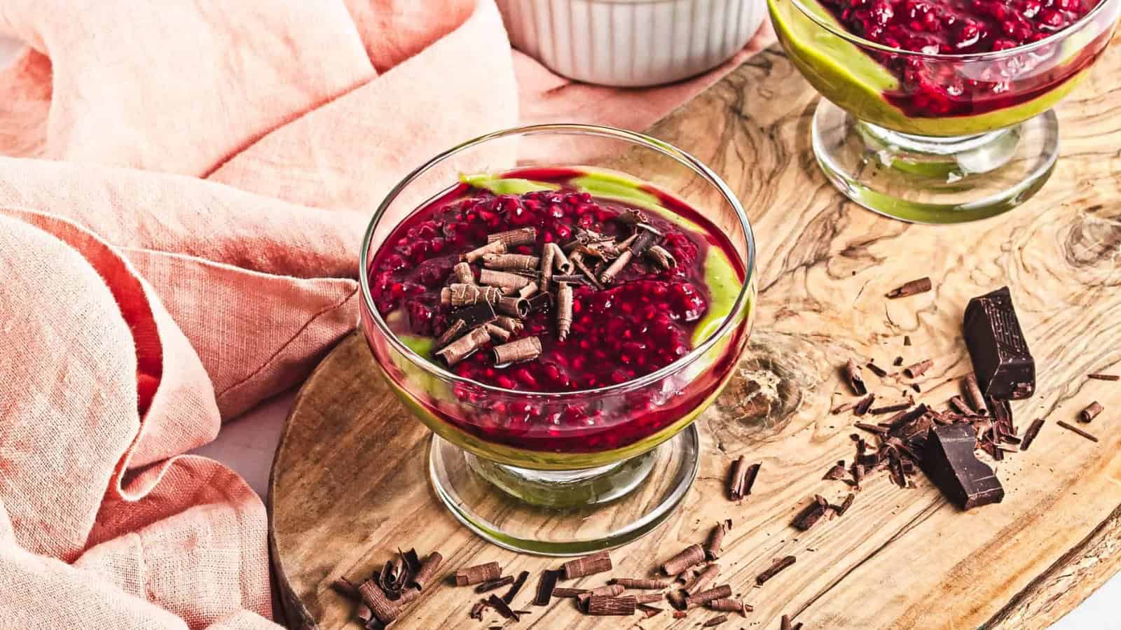 Glass dessert dish with layered green and red mousse, topped with chocolate shavings on a wooden board.
